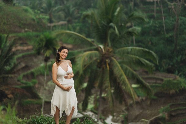 Mujer embarazada joven en vestido blanco con vista a las terrazas de arroz Bali a la luz del sol de la mañana. Armonía con la naturaleza. Concepto de embarazo
. - Foto, Imagen