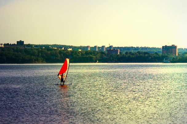 Een eenzame atleet beweegt op een plank met een zeil over het waterige oppervlak. In de verte is een moderne stad, hoge gebouwen. Vroege avond. Zonneverlichting. - Foto, afbeelding