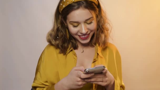 Cute girl in yellow light, laughs, smiles and looks at the camera. A teenager with accessories and a hoop on his head. The girl looks at the phone, then at the camera. The girl uses the application  - Filmati, video