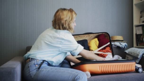 Side view of Travel woman sitting on sofa and packing suitcase getting ready for road trip - Imágenes, Vídeo