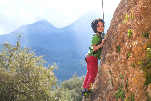 El niño está escalando en un terreno natural. Un niño sube una roca sobre un fondo de montañas. Un hobby extremo. Un niño atlético entrena para ser fuerte. Seguridad en escalada. - Foto, Imagen
