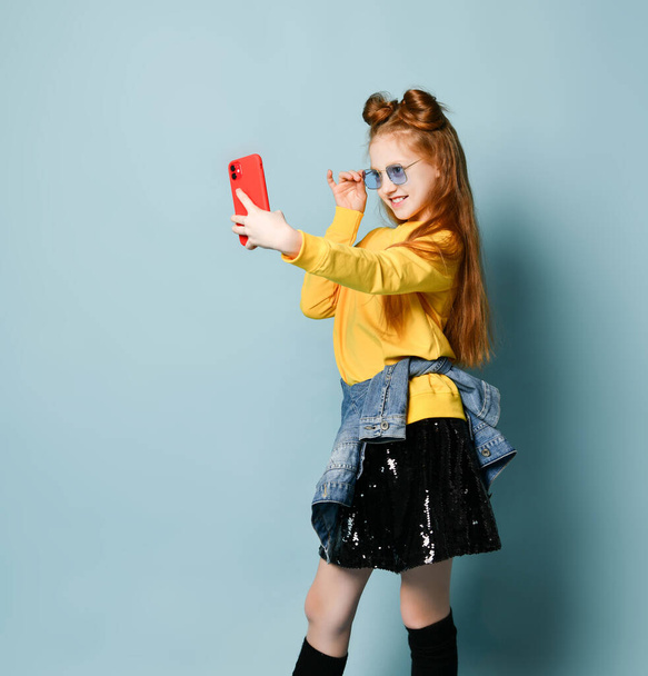 Chica adolescente pelirroja en gafas ahumadas azules, sudadera amarilla y falda negra brillante se está tomando selfie con teléfono inteligente
 - Foto, imagen
