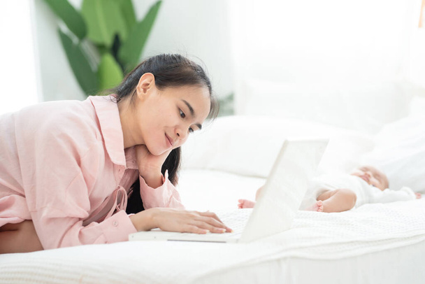 Nuori onnellinen äiti aasialainen nainen työskentelee kannettavan tietokoneen sängyllä huolehtia vastasyntyneen nukkua, työskennellä kotona ja päiväkoti käsite
. - Valokuva, kuva