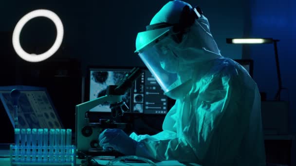 Suojapukuinen tutkija ja tutkimuslaboratoriossa laboratoriolaitteilla työskentelevät naamiot: mikroskoopit, koeputket. Coronavirus SARS-CoV-2 vaara, lääketieteellinen löytö, bakteriologia ja virologia
 - Materiaali, video