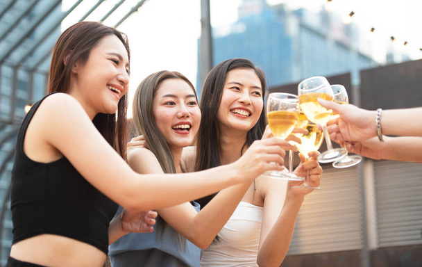 Ομάδα ασιατικών πολλαπλών φύλων κρατώντας ένα ποτήρι κρασί chat μαζί με τους φίλους, ενώ γιορτάζει το πάρτι χορού σε υπαίθριο κλαμπ ταράτσα, τον τρόπο ζωής αναψυχής των νέων έννοια απόλαυση της φιλίας. - Φωτογραφία, εικόνα