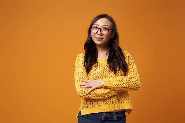 Oriental woman in glasses in sweater with crossed arms in studio - Zdjęcie, obraz