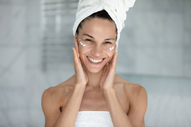 Femme heureuse enveloppée dans une serviette appliquée visage crème toucher la peau
 - Photo, image