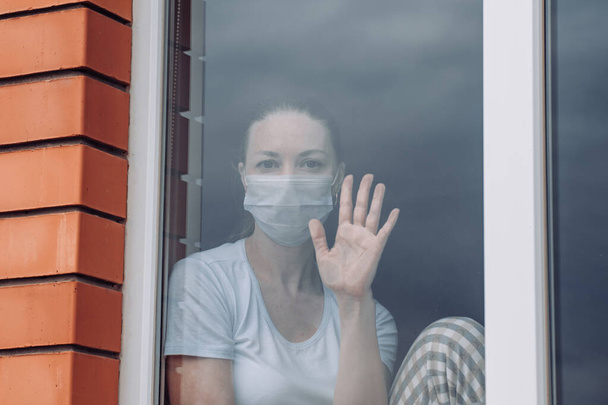 Home quarantine. A Caucasian woman is sitting at window in a medical mask , looking out, her hand on glass, wants to get out. protection against coronavirus infection, pandemics, outbreaks epidemics - Foto, imagen