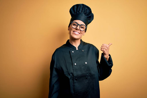 Joven hermosa mujer chef morena con uniforme de cocina y sombrero sobre fondo amarillo sonriendo con la cara feliz mirando y señalando a un lado con el pulgar hacia arriba
. - Foto, Imagen