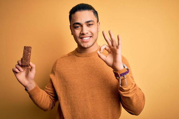 Young brazilian man eating healthy energy bar with protein over isolated yellow background doing ok sign with fingers, excellent symbol - Foto, Imagen