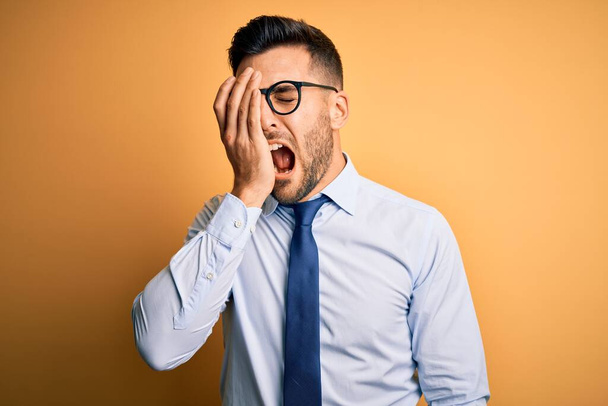 Young handsome businessman wearing tie and glasses standing over yellow background Yawning tired covering half face, eye and mouth with hand. Face hurts in pain. - Photo, Image