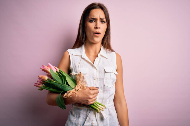 Jeune belle femme brune tenant un bouquet de fleurs de tulipes sur fond rose Au visage choqué, à l'air sceptique et sarcastique, surpris par la bouche ouverte
 - Photo, image