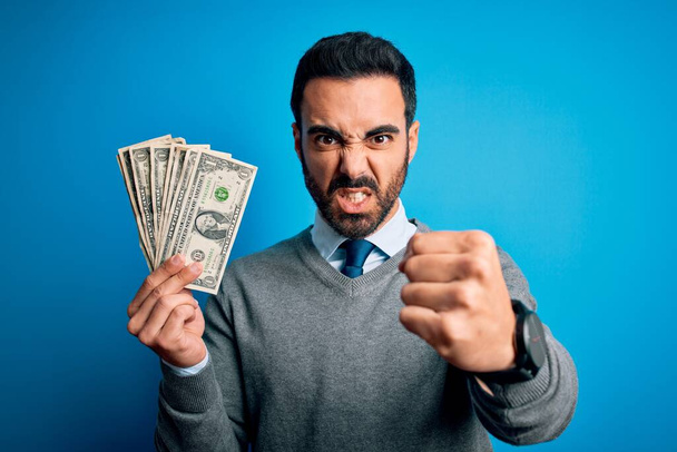 Młody przystojny mężczyzna z brodą trzymający kilka dolarów banknotów na niebieskim tle zirytowany i sfrustrowany krzykiem gniewu, szalony i wrzeszczący podniesioną ręką, koncepcja gniewu - Zdjęcie, obraz