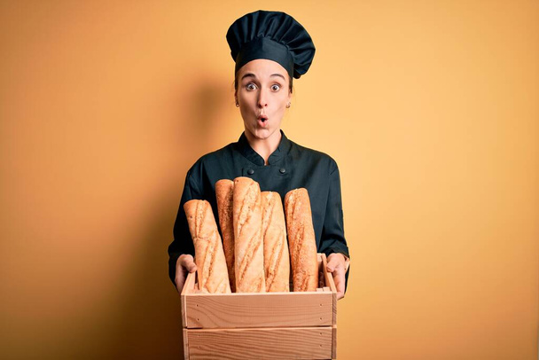 Young beautiful baker woman wearing cooker uniform and hat holding wooden box with bread scared in shock with a surprise face, afraid and excited with fear expression - 写真・画像