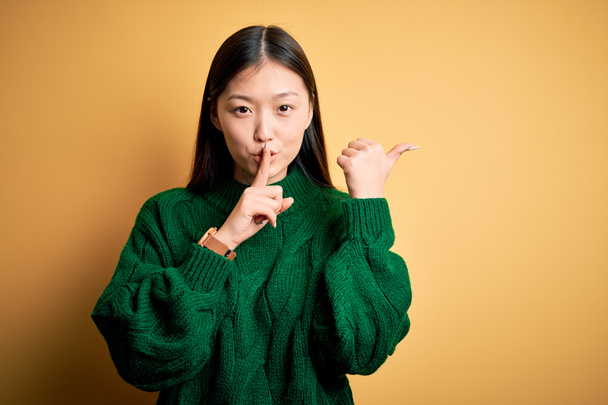 Mladá krásná asijská žena v zeleném zimním svetru přes žluté izolované pozadí žádá, aby byl zticha s prstem na rtech ukazuje s rukou na stranu. Mlčení a tajný koncept. - Fotografie, Obrázek