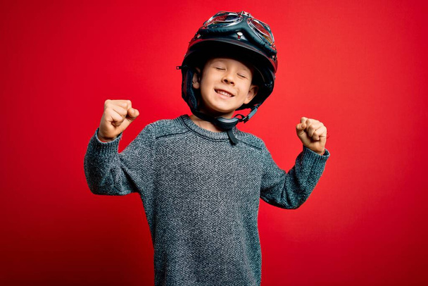Młody, biały dzieciak noszący zabytkowy kask motocyklowy i google na czerwonym tle bardzo szczęśliwy i podekscytowany wykonując gest zwycięzcy z podniesionymi ramionami, uśmiechnięty i krzyczący o sukces. Koncepcja uroczystości. - Zdjęcie, obraz