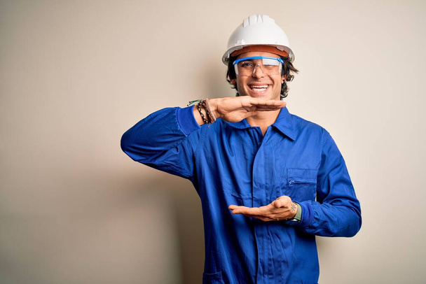 Young constructor man wearing uniform and security helmet over isolated white background gesturing with hands showing big and large size sign, measure symbol. Smiling looking at the camera. Measuring concept. - Photo, Image