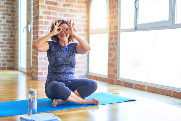Middle age beautiful sportswoman wearing sportswear sitting on mat practicing yoga at home doing ok gesture like binoculars sticking tongue out, eyes looking through fingers. Crazy expression. - Foto, imagen
