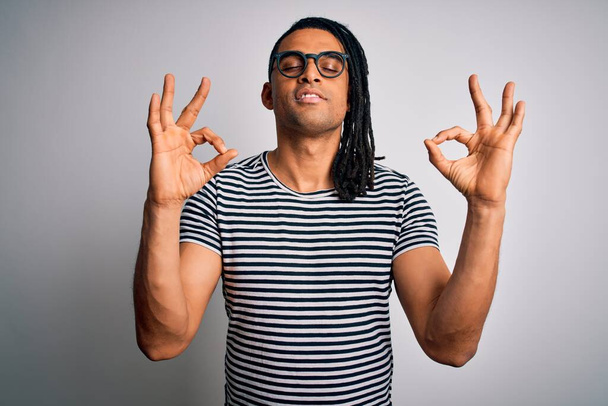 Ein junger gutaussehender afrikanisch-amerikanischer Mann mit Dreadlocks, gestreiftem T-Shirt und Brille entspannt sich und lächelt mit geschlossenen Augen und macht Meditationsgesten mit den Fingern. Yoga-Konzept. - Foto, Bild