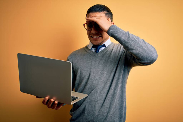 Jonge Braziliaanse zakenman die werkt met een laptop die over een geïsoleerde gele achtergrond staat gestrest met hand op hoofd, geschokt door schaamte en verrassingsgezicht, boos en gefrustreerd. Angst en overstuur voor fouten. - Foto, afbeelding