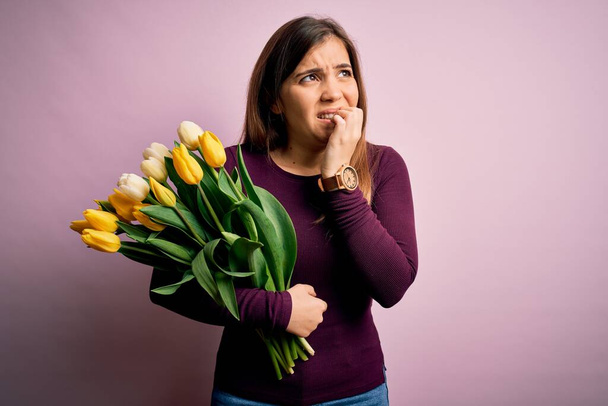 Giovane donna bionda che tiene romantico bouquet di fiori tulipani gialli su sfondo rosa cercando stressato e nervoso con le mani in bocca mordere le unghie. Problema di ansia
. - Foto, immagini