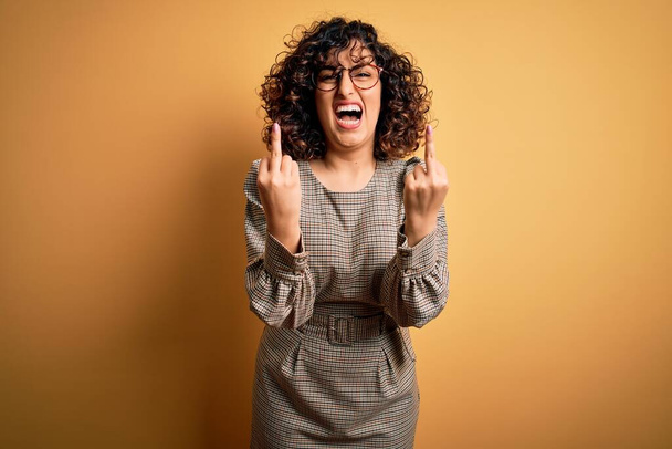 Beautiful arab business woman wearing dress and glasses standing over yellow background Showing middle finger doing fuck you bad expression, provocation and rude attitude. Screaming excited - 写真・画像