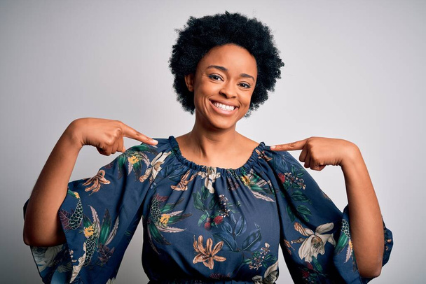 Junge schöne afroamerikanische Afro-Frau mit lockigem Haar trägt lässiges florales Kleid und sieht selbstbewusst aus, mit einem Lächeln im Gesicht, und zeigt mit den Fingern stolz und glücklich. - Foto, Bild