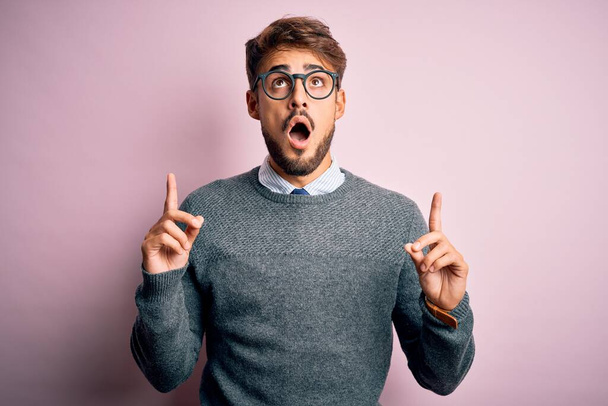 Young handsome man with beard wearing glasses and sweater standing over pink background amazed and surprised looking up and pointing with fingers and raised arms. - Foto, Imagem