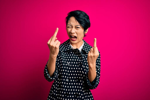 Young beautiful asian girl wearing casual jacket standing over isolated pink background Showing middle finger doing fuck you bad expression, provocation and rude attitude. Screaming excited - Foto, Bild