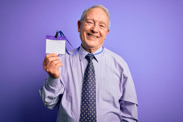 Hombre de negocios de pelo gris mayor sosteniendo la etiqueta de identificación sobre el fondo púrpura con una cara feliz de pie y sonriendo con una sonrisa confiada mostrando los dientes
 - Foto, imagen