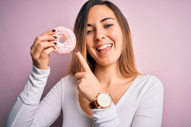 Young beautiful blonde woman with blue eyes holding pink doughnut over isolated background very happy pointing with hand and finger - Photo, Image