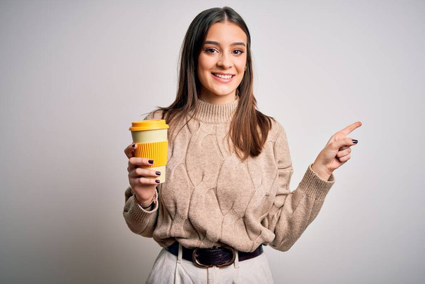 Молодая красивая брюнетка женщина пьет кофе на изолированном белом фоне очень счастливы указывая рукой и пальцем в сторону
 - Фото, изображение