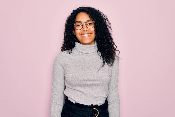 ピンクの背景にタートルネックのセーターと眼鏡を身に着けている若いアフリカ系アメリカ人女性は、顔に幸せとクールな笑顔で。運のいい人. - 写真・画像