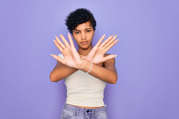 Joven hermosa afroamericana afro mujer con camiseta casual sobre fondo púrpura expresión de rechazo cruzando brazos y palmas haciendo signo negativo, cara enojada
 - Foto, imagen