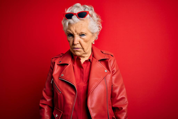 Senior mooie grijs-harige vrouw draagt casual rode jas en zonnebril sceptisch en nerveus, fronsen boos als gevolg van problemen. Negatief persoon. - Foto, afbeelding