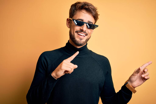 Young man wearing thug life fanny sunglasses standing over isolated yellow background smiling and looking at the camera pointing with two hands and fingers to the side. - Foto, immagini