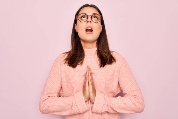 Fiatal gyönyörű barna nő visel alkalmi pulóver és szemüveg felett rózsaszín háttér koldulás és imádkozás kéz együtt remény kifejezés az arcon nagyon érzelmes és aggódik. Könyörgök.. - Fotó, kép