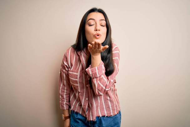 Young brunette woman wearing casual striped shirt over isolated background looking at the camera blowing a kiss with hand on air being lovely and sexy. Love expression. - Foto, afbeelding