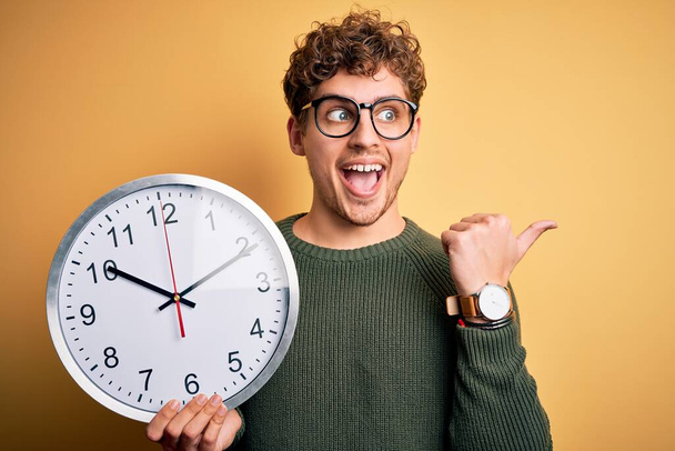 Young blond man with curly hair wearing glasses holding big clock over yellow background pointing and showing with thumb up to the side with happy face smiling - Foto, Bild
