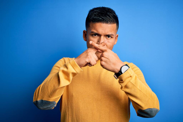 Mladý pohledný latinsky muž na sobě žlutý ležérní svetr přes izolované modré pozadí odmítání výraz křížení prsty dělá negativní znamení - Fotografie, Obrázek