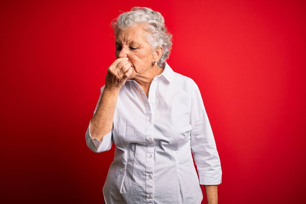 Старшая красивая женщина в элегантной рубашке, стоящей на изолированном красном фоне, пахнет чем-то вонючим и отвратительным, невыносимым запахом, затаив дыхание пальцами на носу. Плохой запах
 - Фото, изображение