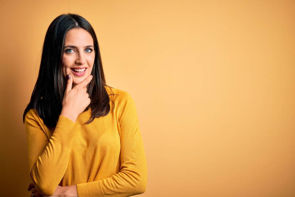 Young brunette woman with blue eyes wearing casual sweater over yellow background looking confident at the camera smiling with crossed arms and hand raised on chin. Thinking positive. - Φωτογραφία, εικόνα