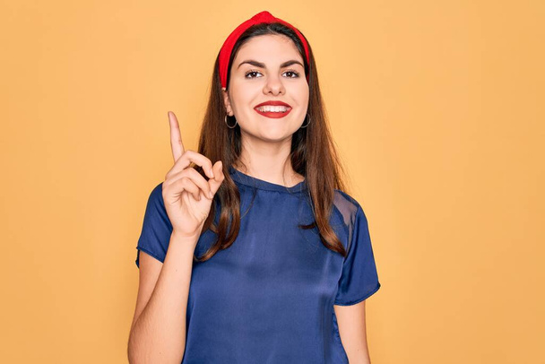 Young beautiful brunette woman wearing red lips over yellow background showing and pointing up with finger number one while smiling confident and happy. - Photo, Image