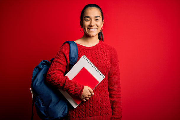 Jovem bela estudante asiática mulher vestindo mochila em pé sobre isolado fundo vermelho com um rosto feliz de pé e sorrindo com um sorriso confiante mostrando dentes
 - Foto, Imagem