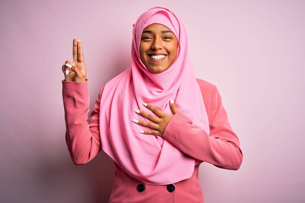 Junge afroamerikanische Afro-Frau trägt muslimischen Hijab vor isoliertem rosa Hintergrund, lächelt und schwört mit der Hand auf Brust und erhobenen Fingern und leistet einen Treueschwur - Foto, Bild