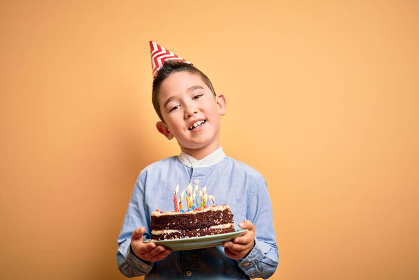 Uroczy maluch noszący czapkę urodzinową trzymający tort ze świeczkami stojącymi nad odizolowanym żółtym tłem - Zdjęcie, obraz