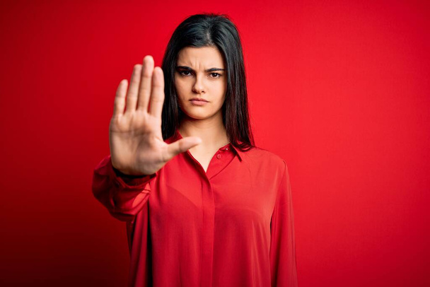 赤い背景の上に立つカジュアルなシャツを着た若い美しいブルネットの女性は、手の手のひらで歌うのをやめます。顔に負と深刻なジェスチャーで警告式. - 写真・画像