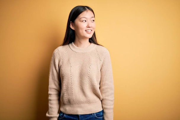 Fiatal gyönyörű ázsiai nő visel alkalmi pulóver sárga elszigetelt háttér néz oldalra mosollyal az arcon, természetes kifejezés. Nevetve magabiztos. - Fotó, kép