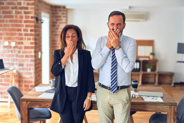 Two middle age business workers standing working together in a meeting at the office bored yawning tired covering mouth with hand. Restless and sleepiness. - Photo, Image