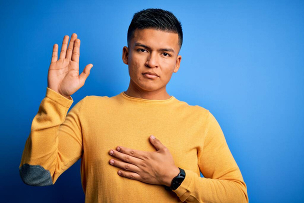 Joven hombre latino guapo usando suéter casual amarillo sobre fondo azul aislado jurando con la mano en el pecho y la palma abierta, haciendo un juramento de lealtad promesa
 - Foto, imagen
